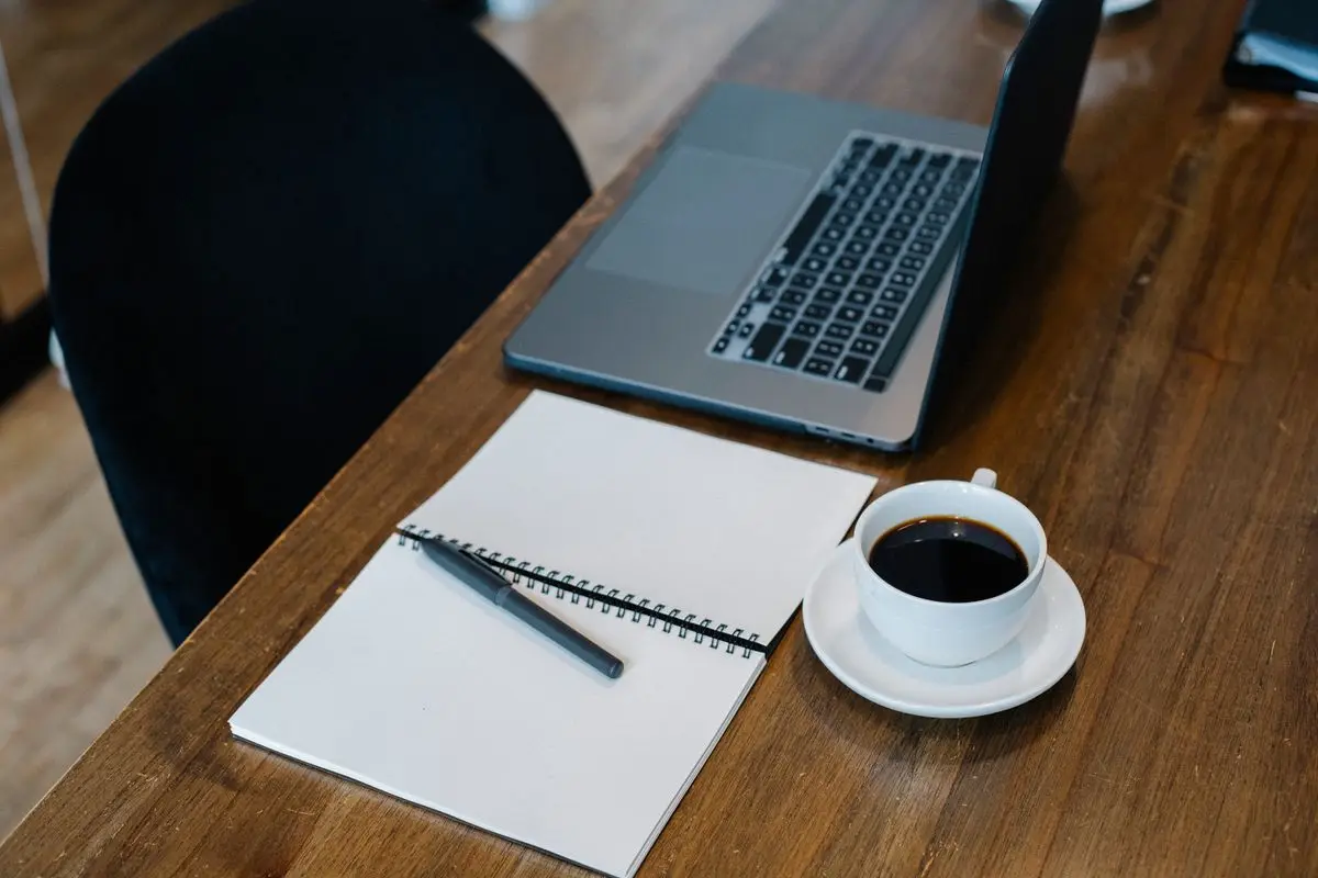 A laptop, blank notebook, pen and cup of coffee on a wooden desk